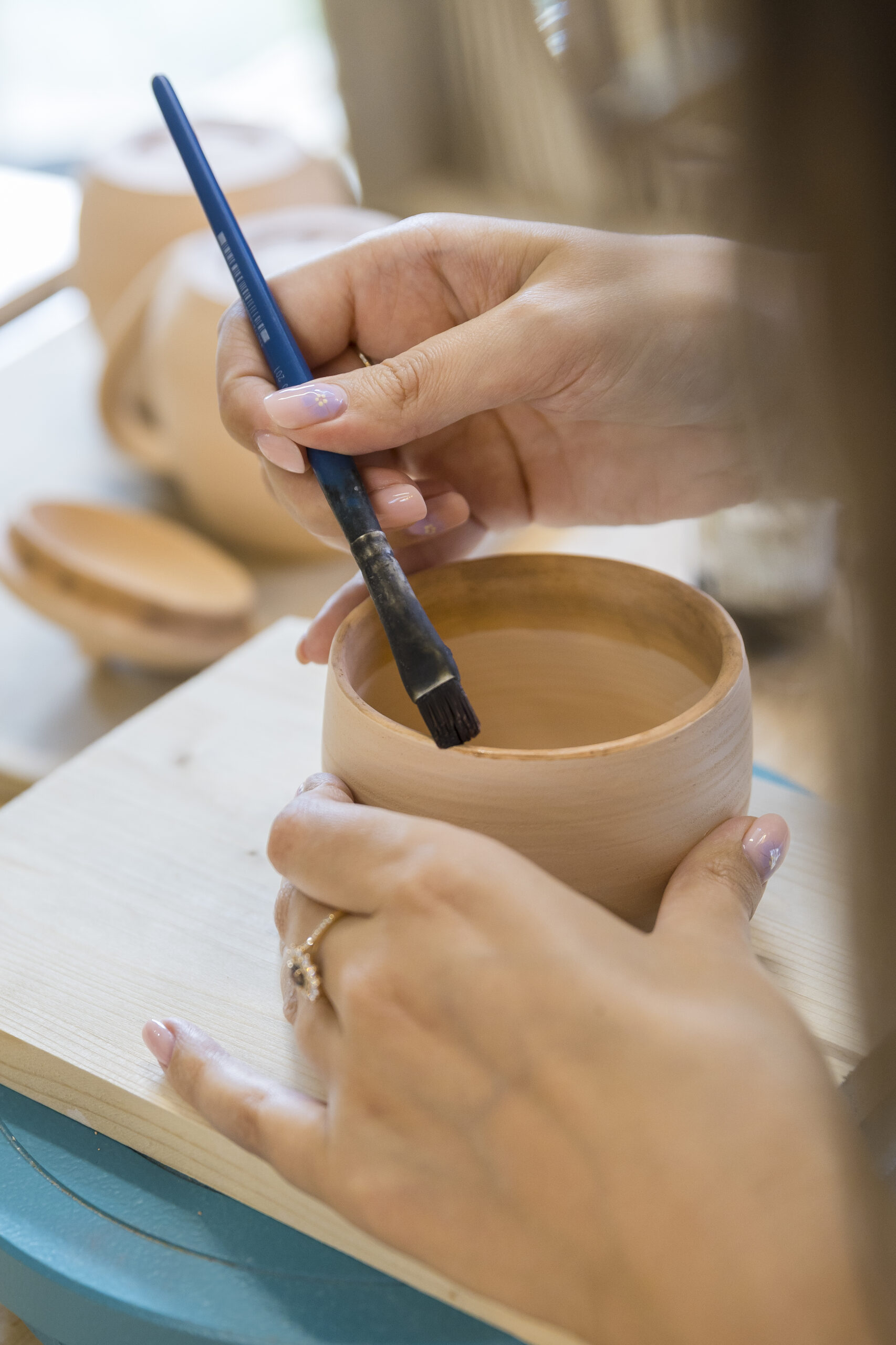 Application de cire avant l'émaillage. Photographie de Emmanuelle Bayart Photographie de deux mains. L'une tient une tasse, l'autre un pinceau qui peint sur le bord de la tasse.